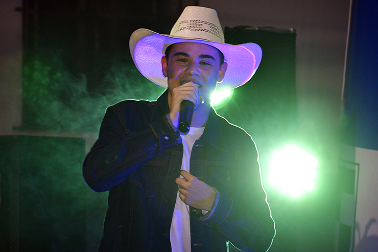 Elifas Magalhães é um homem branco, está ao centro da imagem com refletores ao fundo. Ele segura um microfone e usa um chapéu branco e está sorrindo enquanto se apresenta no Show de Talentos.