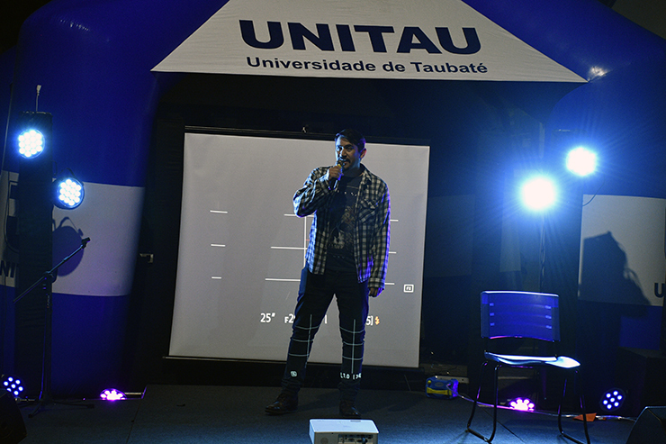 Bertino é um homem de cabelos e barbas escuras e aparece em pé ao centro da imagem, com refletores ao fundo. Ele segura um microfone enquanto se apresenta no Show de Talentos.