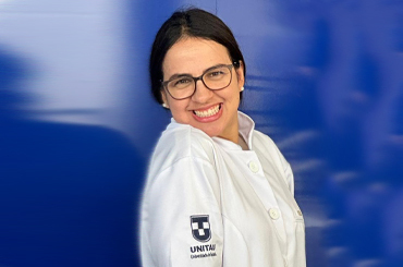Soraia é uma mulher branca e usa óculos. Ela posa sorridente com o jaleco bordado com o símbolo da Universidade de Taubaté em um fundo azul. 